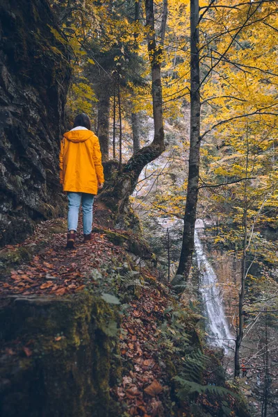 Frau spaziert durch herbstlichen Waldweg im gelben Regenmantel Wasserfall im Hintergrund — Stockfoto