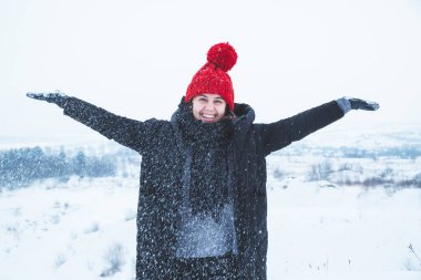 Gülümseyen, kırmızı şapkalı, dışarıda karla oynayan yetişkin bir kadın.