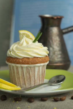 Tatlı limon cupcakes ve sıcak kahve ile güzel kahvaltı zamanı, kaşık ve kavrulmuş kahve çekirdekleri ile plaka, peynir kremi, limon dilim, keten masa örtüsü mavi yemek kitabı arka plan biberiye dalı, arka ve kopya alanı bulanık cezve ile