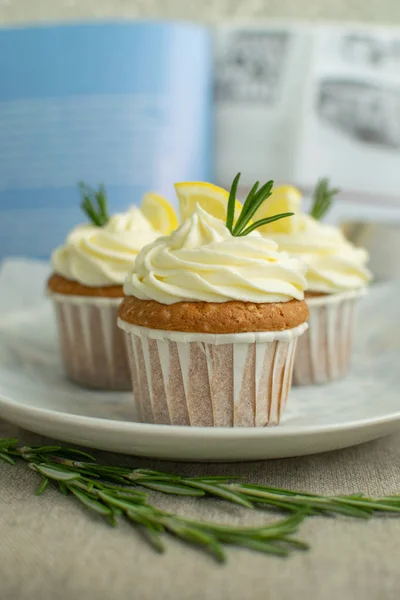 Bakery lemon cupcakes with cheese cream, slice of lemon, decorated by rosemary at blue cookbook background at linen tablecloth on plate, selective focus, close up, copy space.