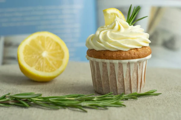 Homemade bakery lemon cupcakes with cheese cream, slice of lemon, branch of rosemary at blue cookbook background at linen tablecloth, selective focus, close up, copy space, from the top.