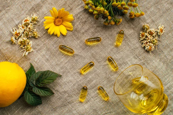 Layout with fish oil capsules, tansy, dry chamomile flowers, lemon, calendula, glass cup, raspberry leaf on linen fabric at the table, copy space — Stock Photo, Image