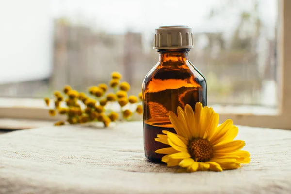 Herbal tincture in pharmacy bottle and yellow flower of calendula with tansy at background at linen textile, alternative medicine