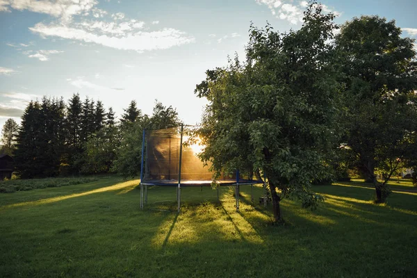 Nyílt gyermek ugrás trambulin a napon a zöld fű az udvaron a finn ház fákkal, nyári este — Stock Fotó