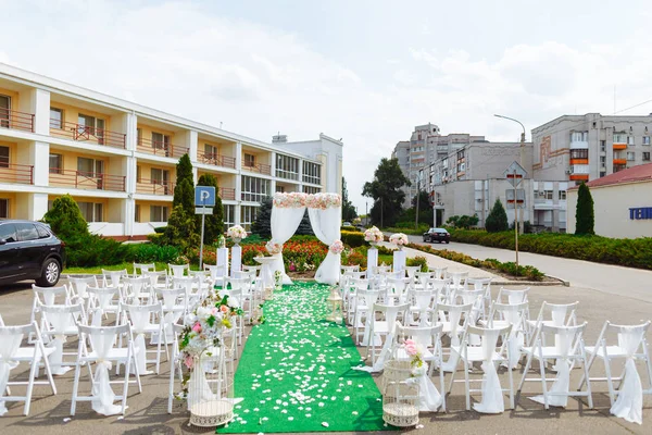 Beautiful decoration of the wedding ceremony. White wooden chairs for guests on either side of the walkway, which leads to festive arch decorated with flowers