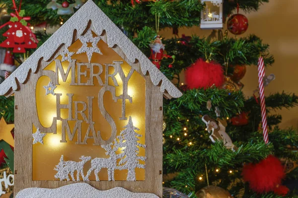 Wooden crib design with Merry Christmas message before lit tree. A wood carved illuminated crib with a winter scene against a decorated artificial Christmas tree background.