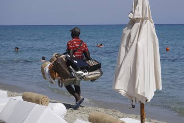 Chaniotis, Greece - July 08 2017: Vendor with counterfeit products on the beach.Street vendor selling popular brands replicas bags by a Greek beach shore on a sunny summer day. clipart