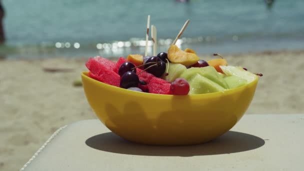 ビーチで木のつまようじと夏の新鮮なフルーツサラダ 夏の日にぼやけた海の背景にメロン スイカ サクランボ 赤ブドウ アプリコットのような刻んだ新鮮な果物の黄色いボウル — ストック動画