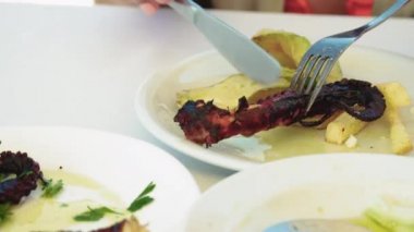 Yunan taverna restoran ahtapot closeup hizmet vermektedir. Dilimlenmiş domates, salatalık ve kabak ile beyaz bir tabak üzerinde gevrek kömürleşmiş ahtapot tentacle hizmet çatal ve bıçak ile el, bir taverna masada.