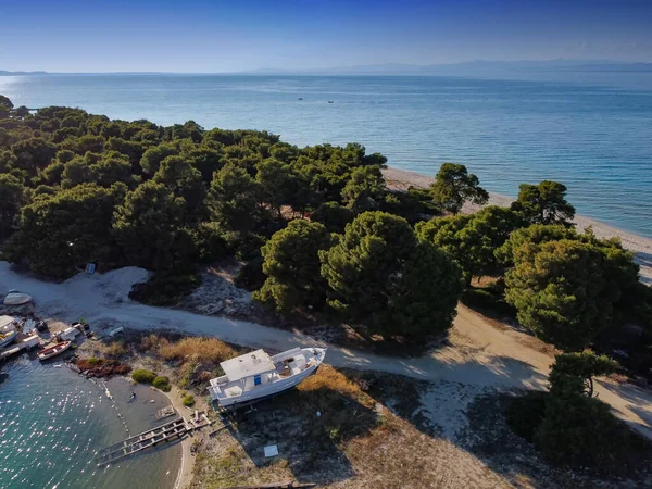 Mediterranean Greek landscape coastal drone shot with caique boat on shore. Aerial top view of Glarokavos lagoon at Kassandra Chalkidiki peninsula above shoreline with green plantation & calm waters.