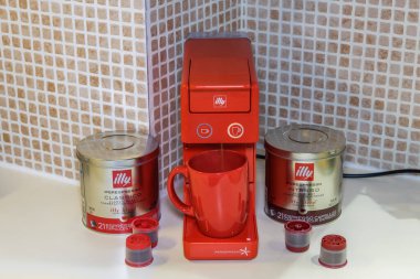 Tek servis kapsüllü espresso yapmak için kullanılan otomatik Illy Iperespresso makinesi. Kırmızı elektrikli kahve makinesinin etrafında bavul ve şirket logosu olan kapsüller, fincanda sıcak damlayan kahve yapmak için..