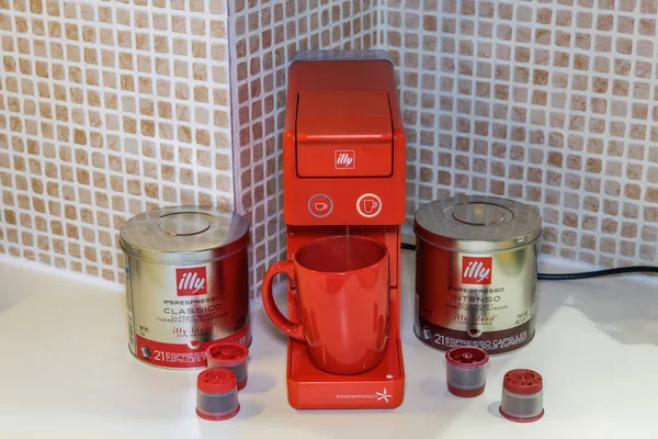Tek servis kapsüllü espresso yapmak için kullanılan otomatik Illy Iperespresso makinesi. Kırmızı elektrikli kahve makinesinin etrafında bavul ve şirket logosu olan kapsüller, fincanda sıcak damlayan kahve yapmak için..
