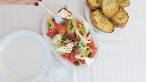 女性はフェタチーズとギリシャのサラダを提供しています 玉ねぎ トマト パセリ キュウリ スプーン フォークトップビューで提供しています居酒屋テーブルの白い皿のXoriatikiの閉鎖 パンのスライスの横にある — ストック動画