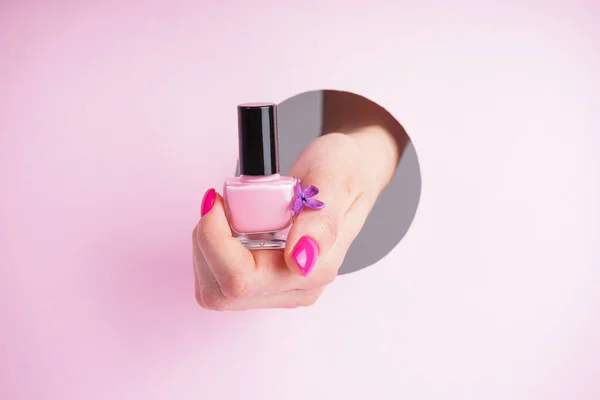 Beautiful manicure close-up. Beautiful hand of a young woman holds a tube of nail polish and a lilac flower on a pink background.