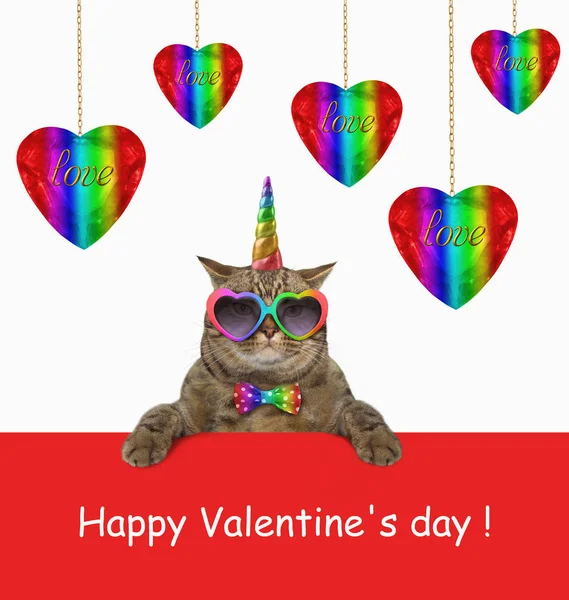The cat unicorn in a bow tie and funny sunglasses is sitting next to color shiny hearts hanging on gold plated chains. Happy Valentines Day.