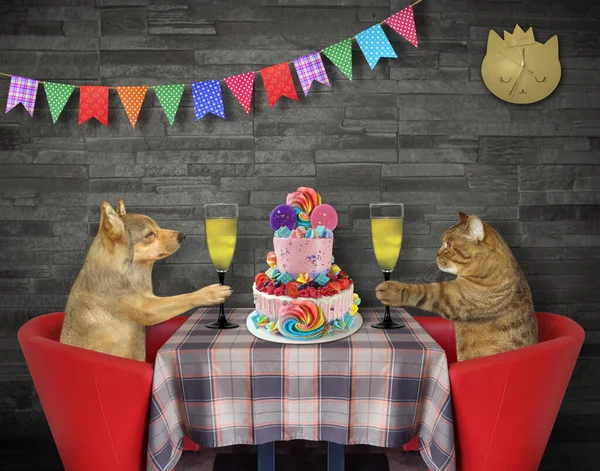 A cat with a dog together are eating a holiday two tiered cake and drinking wine at a table in a restaurant.