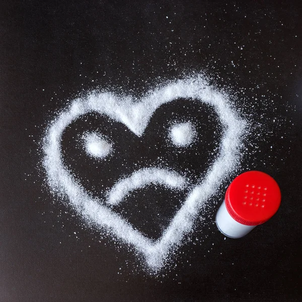 盐散落在黑色的表面上 画的心与悲伤的脸 对健康的危害从过量盐和糖的消耗量 — 图库照片
