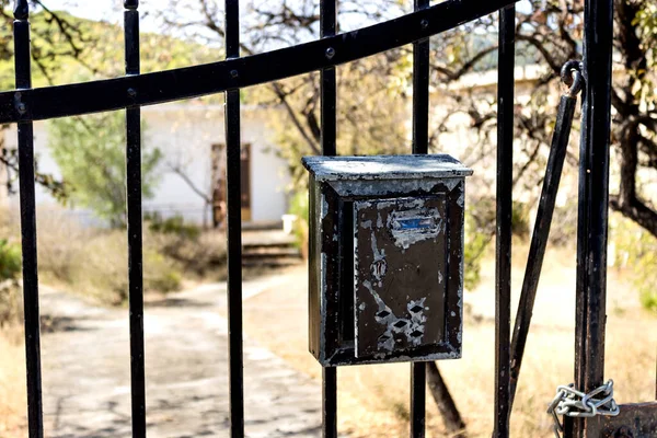 A barred gate with an old mailbox, against the background of a small white house and an overgrown garden. Selective focus.
