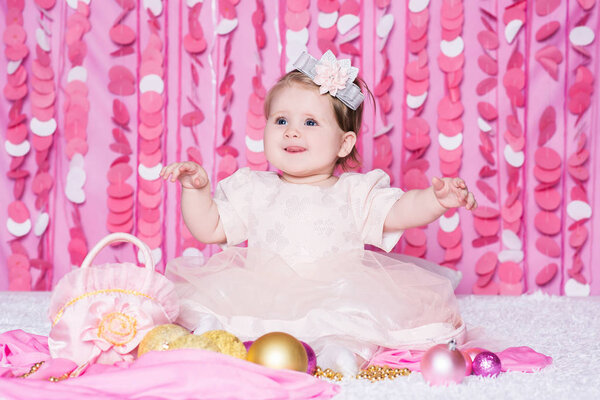 Симпатичная маленькая девочка сидит на мягком одеяле в розовом платье с рождественскими шарами, праздничный подарок и сумка, в праздничном розовом интерьере. Новогодние и рождественские праздники
.