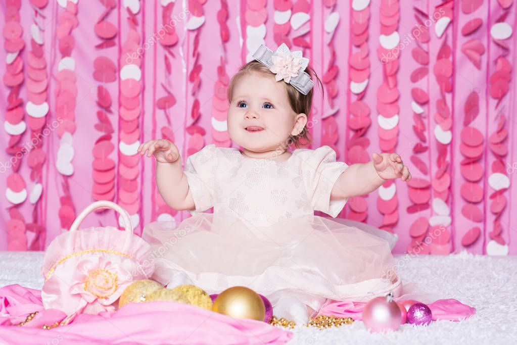 Cute little baby girl sits on a soft blanket in a pink dress with a Christmas balls, party gift and handbag, in a festive pink interior. New Year and Christmas holidays.
