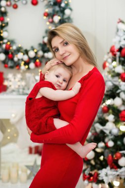 Mutlu Genç Anne üzerinde ellerini onun şirin bebek kız Noel ve yeni yıl tatilleri üzerinde tutar. Çocuk ile anne Noel ağacı ve bulanık şenlikli ışıkları ile dekore edilmiş oda kırmızı elbiseler