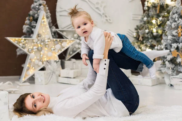 Şirin bebek kızı ile Noel ve yeni yıl tatilleri üzerinde oynarken mutlu genç anne. Çocuk ile anne Noel ağacı ve bulanık şenlikli ışıkları ile dekore edilmiş Oda