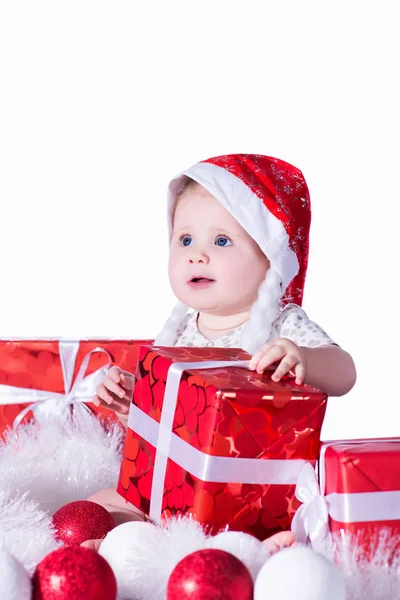 Küçük şirin bebek kız, Noel Baba, kırmızı bir şapka kırmızı ve beyaz Noel topları, kırmızı hediye kutuları ve çelenk çevrili, beyaz arka plan üzerinde izole oturuyor. Yeni yıl ve Noel tatilleri.
