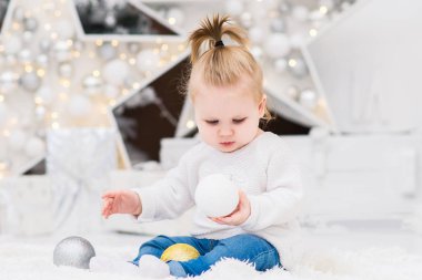 Küçük şirin bebek kız Noel ağaçları, ışıklar, çelenk, Noel topları ve hediye kutuları bir arka plan üzerinde beyaz bir kazak oturuyor. Yeni yıl ve Noel tatilleri.