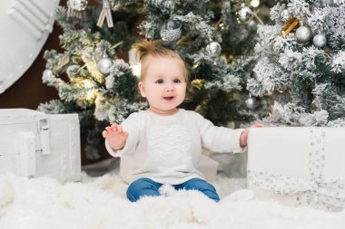 Noel ağaçları, ışıklar, çelenk ve hediye kutuları bir arka plan üzerinde beyaz bir kazak şirin bebek kız gülümsüyor. Yeni yıl ve Noel tatilleri.
