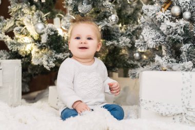 Noel ağaçları, ışıklar, çelenk ve hediye kutuları bir arka plan üzerinde beyaz bir kazak şirin bebek kız gülümsüyor. Yeni yıl ve Noel tatilleri.