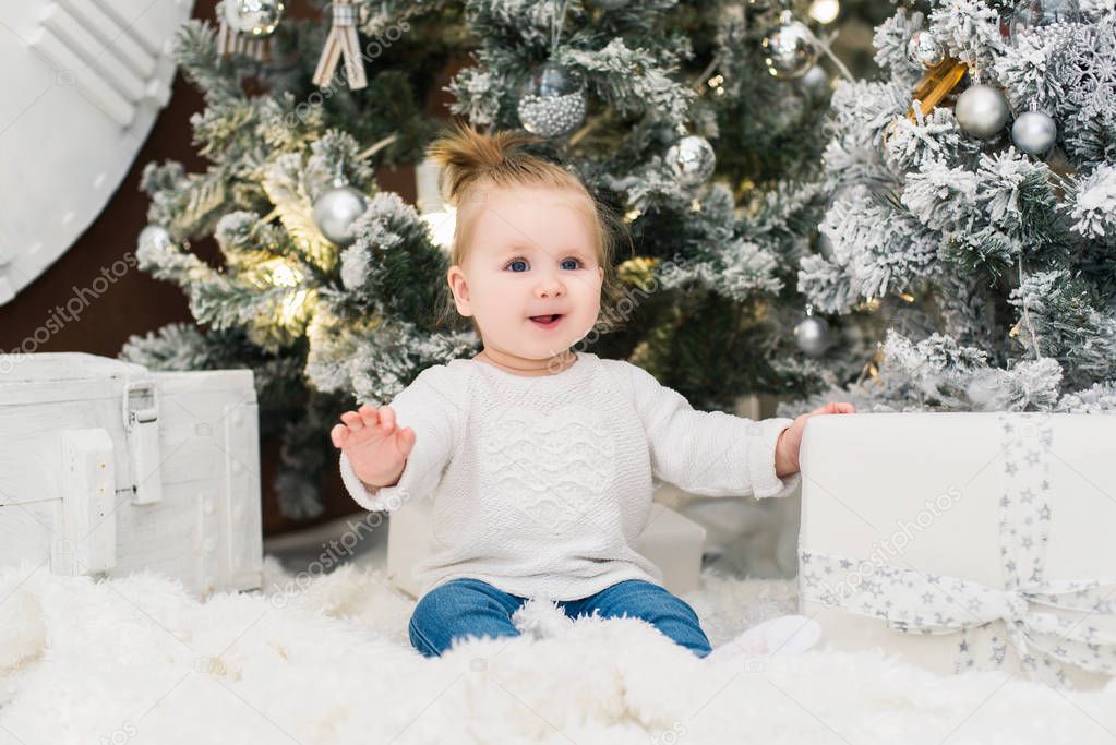 Smiling cute baby girl in a white sweater on a background of Christmas trees, lights, garlands and gift boxes. New Year and Christmas holidays.