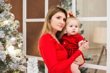 Mutlu Genç Anne üzerinde ellerini onun şirin bebek kız Noel ve yeni yıl tatilleri üzerinde tutar. Çocuk ile anne Noel ağacı ve bulanık şenlikli ışıkları ile dekore edilmiş oda kırmızı elbiseler