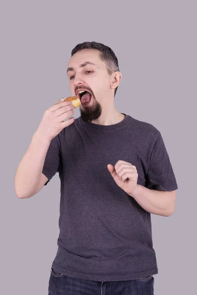 Homem com barba comendo um pão sobre fundo cinza — Fotografia de Stock