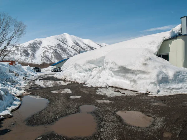 Buildings covered with snow, a spring road with puddles and a view of the snow-covered hills on a sunny spring day