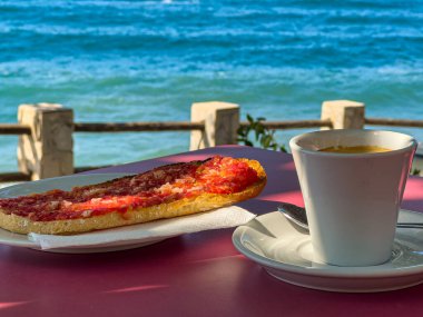 Bir espressonun yatay fotoğrafı ve Akdeniz kıyısındaki bir masada domates ve yağlı tost. Ağaçtan gölgeli doğal ışık ve metin için geniş alan.