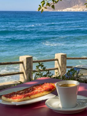 Açık hava masasında espresso kahve, domatesli tost, yağ ve tuzlu kahvaltı fotoğrafı. Arka planda Akdeniz, kıyı manzarası ve yapraklı dallar var..