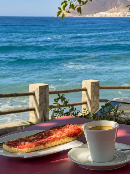 Açık hava masasında espresso kahve, domatesli tost, yağ ve tuzlu kahvaltı fotoğrafı. Arka planda Akdeniz, kıyı manzarası ve yapraklı dallar var..
