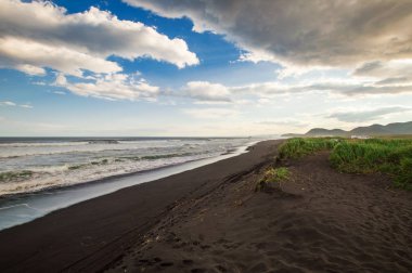 Halaktyr plaj. Kamçatka. Rusya Federasyonu. Karanlık neredeyse siyah renkli kum beach Pasifik Okyanusu. Taş dağları ve sarı çim bir arka plan üzerinde vardır. Hafif mavi gökyüzü.