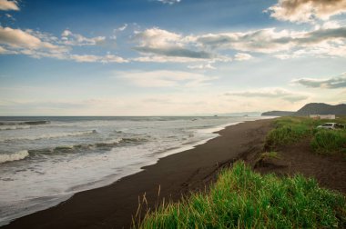 Halaktyr plaj. Kamçatka. Rusya Federasyonu. Karanlık neredeyse siyah renkli kum beach Pasifik Okyanusu. Taş dağları ve sarı çim bir arka plan üzerinde vardır. Hafif mavi gökyüzü