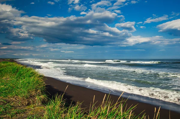 Halaktyr beach. Kamtjatka. Ryska federationen. Mörk nästan svart färg sand stranden av Stilla havet. Sten berg och gula gräset är på en bakgrund. Ljus blå himmel — Stockfoto