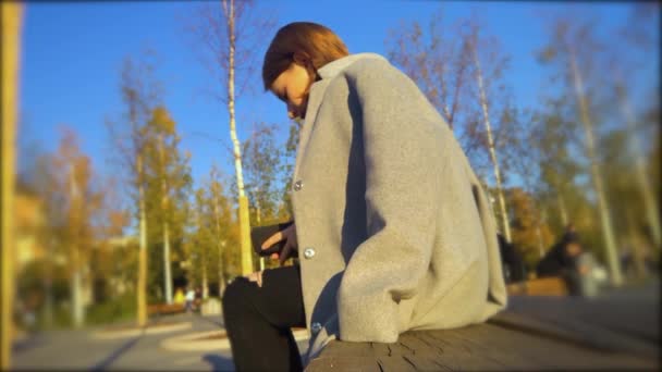 Dívka v plášti sedí v parku za slunečného počasí, směje se a staví na brýle — Stock video