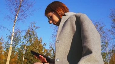 Gri bir ceket ve güneş gözlüğü genç kız güneşli bir günde onu Smartphone bir şey arıyor