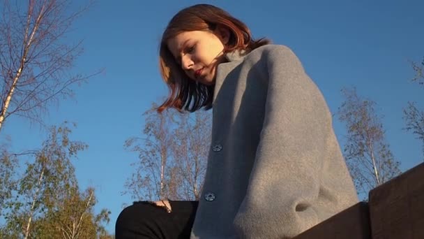 Teen ragazza in un cappotto grigio si siede nel parco e controlla il telefono in una giornata di sole — Video Stock