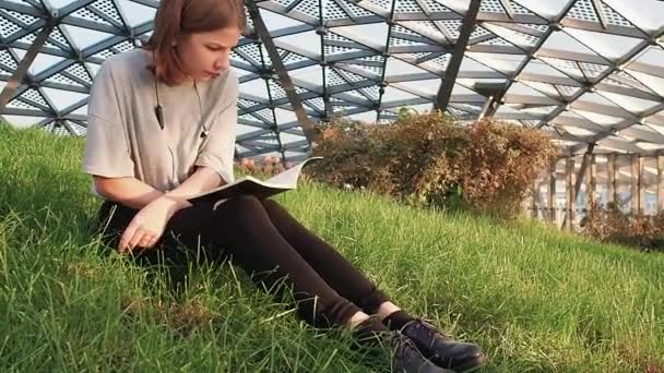 Chica adolescente con una camisa gris sentada en la hierba en el parque y leyendo un cuaderno con notas de conferencia en un día soleado — Vídeos de Stock