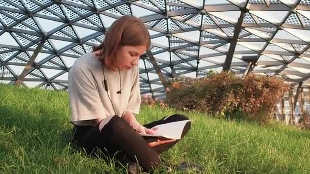 Девочка-подросток в серой рубашке сидит на траве в парке и смотрит вокруг и читает тетрадь с конспектами лекций в солнечный день — стоковое видео
