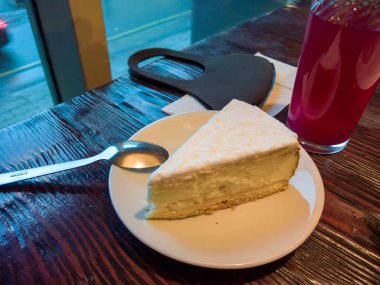 Bir dilim cheesecake kekini çay kaşığıyla birlikte bir çay tabağının üzerinde, ahşap bir masanın üzerinde. Yanında koruyucu bir antiviral maske var. Arka planda caddesi olan bir pencere var..