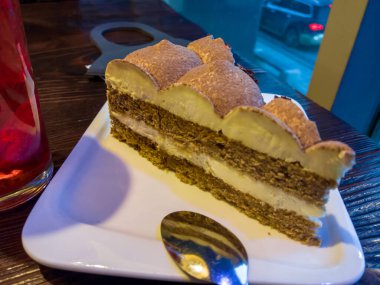 Bir parça tiramisu keki kaba ahşap bir masanın üzerinde beyaz bir fincan tabağı üzerinde. Onun yanında bir bardak çilekli komposto var. Onun yanında Japonya 'dan gelen siyah bir tıbbi maske var. Caddedeki bir pencerenin arkasında..