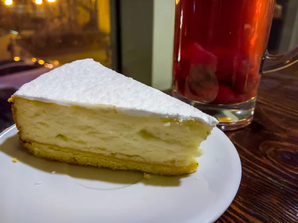 白いソーサーの上にチーズケーキをのせる大きな木のテーブルの上には 通りの背景とイチゴのコンポートのガラスにはっきりとした質感がある — ストック写真