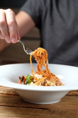 Spagetti bir çatalla bir plaka üzerinde tutan elini. Domates soslu makarna, kurutulmuş domates, parmesan ve fesleğen ahşap arka plan üzerinde