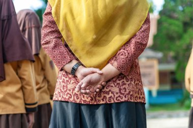 Elleri arkada duran, geleneksel kırmızı batik giyen, kültürü, sabrı ve zarafeti simgeleyen birinin yakın çekimi.